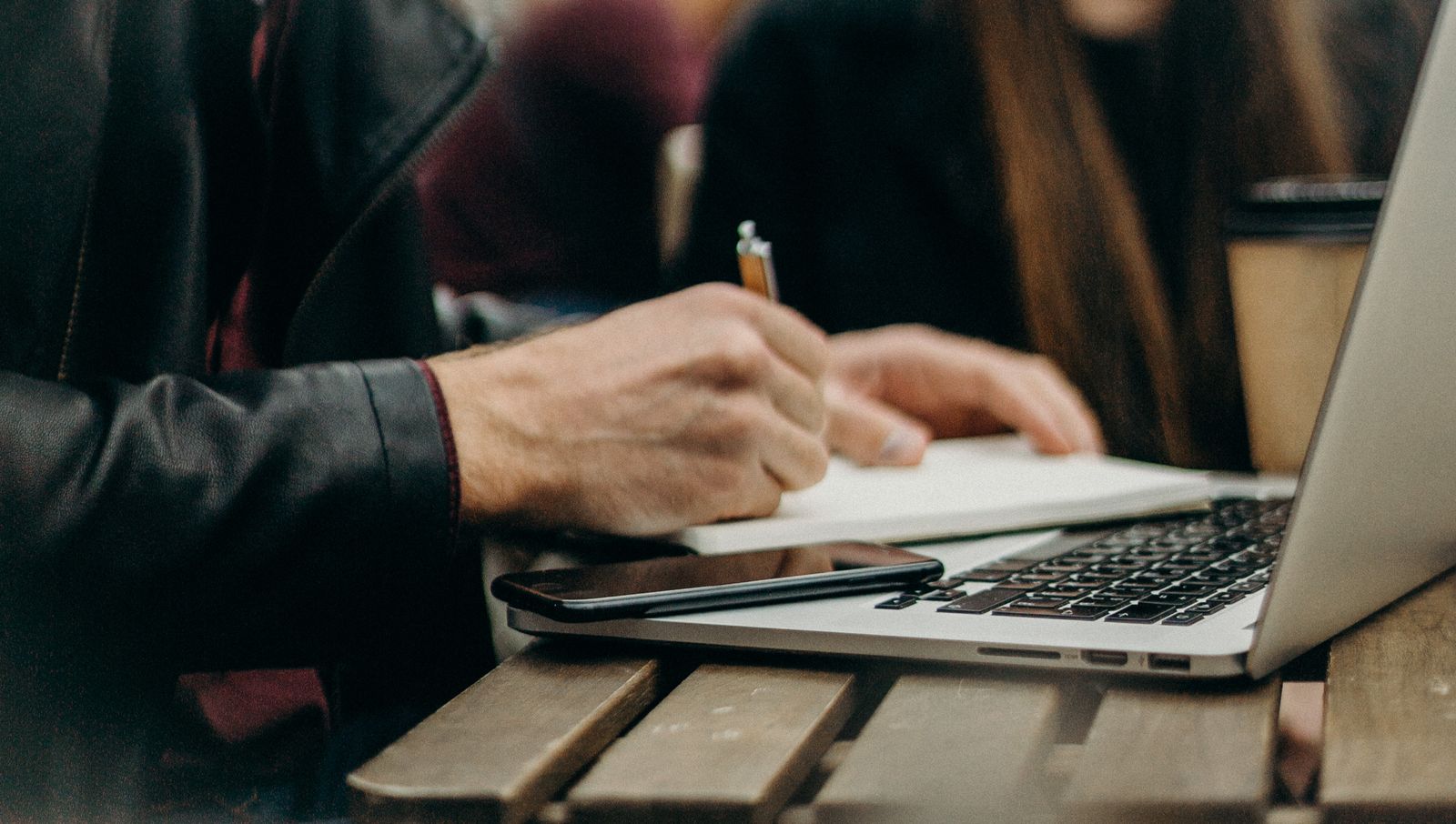 Eine Person in Lederjacke schreibt an einem Holztisch in ein Notizbuch, daneben Laptop und Telefon, im Hintergrund unscharfe Personen.